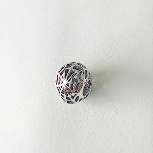 May include: A silver charm with a geometric design. The charm is spherical with a web-like pattern of lines and shapes. The charm has a dark interior and is set against a white background.