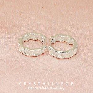 May include: A pair of silver hoop earrings with a textured, chain-like pattern. The earrings are circular and made of metal. The background is a pale pink fabric. The text "CRYSTALINEGB Handcrafted Jewellery" is visible at the bottom.