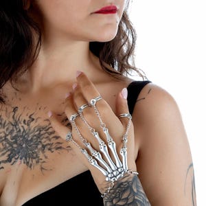 May include: Silver-toned skeleton hand bracelet with rings. The bracelet features a detailed design of a skeletal hand, with rings connected by chains to the finger bones. The jewelry is worn on the hand and wrist, creating a unique and gothic aesthetic.