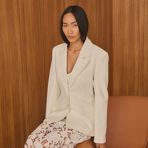 May include: A cream-colored blazer and matching top paired with white lace pants. The blazer has a classic notched lapel and a single-button closure. The pants feature a floral lace pattern. The outfit is worn with a brown chair.