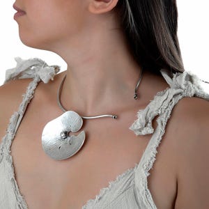 May include: A silver necklace featuring a large, textured pendant with an unusual, organic form. The pendant has a hammered finish and a small, round accent. The necklace is worn around the neck.