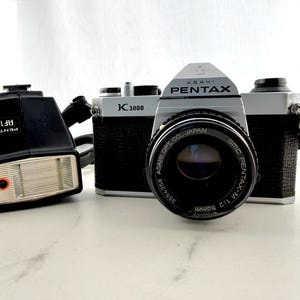 May include: A silver and black Asahi Pentax K1000 35mm film camera with an SMC Pentax-M 1:2 50mm lens attached. Beside the camera sits a black Pentax AF160 electronic flash unit. Both items feature visible branding and model text on their surfaces.