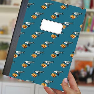Può includere: Un quaderno con una copertina turchese con un motivo ripetuto di gabbiani e patatine fritte. Il quaderno ha un'etichetta bianca vuota e un dorso nero. Il quaderno è tenuto da una persona.
