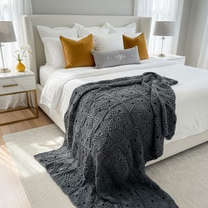May include: A dark gray, crocheted blanket draped over a white bed. The blanket features a geometric pattern with raised textures. The bed is adorned with white and mustard-colored pillows. A neutral rug and bedside tables complete the bedroom setting.