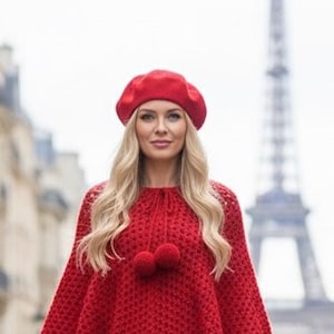 Könnte beinhalten: Ein rotes Barett und ein passender Strickponcho mit Bommelakzenten. Das Outfit ist in einem leuchtenden Rotton gehalten und sorgt für einen stimmigen und stilvollen Look. Der Eiffelturm ist im Hintergrund zu sehen.