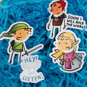 Peut inclure: Ensemble d'autocollants de dessins animés représentant des personnages d'un jeu vidéo fantastique. L'un d'eux montre un personnage avec une épée et le texte "HEY! LISTEN!". Un autre dit "SOON I WILL RULE THE WORL!". Les autocollants sont sur du papier bleu.