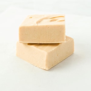 May include: Two square-shaped, light tan bars of soap are stacked on top of each other. The top bar has a textured surface with a swirl design. The soap is sitting on a white surface.