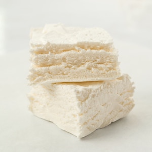 May include: Two pieces of white marshmallow stacked on top of each other. The marshmallow is soft and fluffy, with a slightly grainy texture.