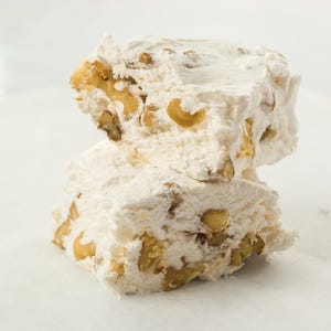 May include: Two stacked pieces of white nougat candy with visible nuts. The nougat has a soft, textured appearance, and the nuts are a light brown color. The candy is on a white surface.