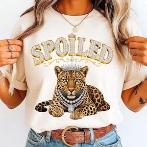 May include: A cream-colored t-shirt featuring a graphic of a leopard wearing a diamond-style crown and a multi-strand diamond necklace. Above the leopard, the word SPOILED is printed in large, gold, diamond-encrusted letters with a crown icon.