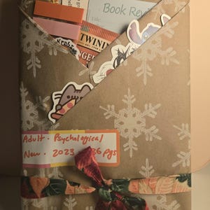 May include: A wrapped book with a brown paper cover, decorated with white snowflakes. The book is tied with a floral ribbon. Stickers and bookmarks peek out from the top. Text on a label reads "Adult - Psychological, New 2023, 36 pgs."