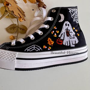 May include: Black high-top canvas sneaker with white laces and a thick white sole. The shoe features embroidered Halloween-themed designs, including a ghost, spiderwebs, candy corn, and a jack-o'-lantern. The text "Beautiful Hourss" is printed on the side.