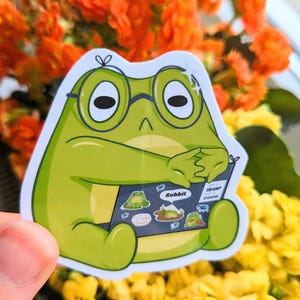 May include: A green frog sticker wearing glasses, holding a book with the words "Rubbit" and "Swamp Code". The sticker has a white border and is set against a backdrop of orange and yellow flowers.