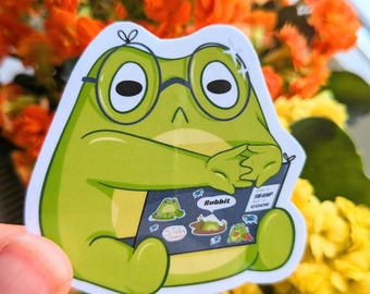 May include: A green frog sticker wearing glasses, holding a book with the words "Rubbit" and "Swamp Code". The sticker has a white border and is set against a backdrop of orange and yellow flowers.