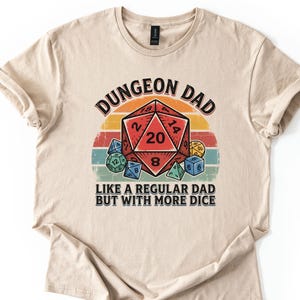 May include: Sand-colored t-shirt with the text "DUNGEON DAD" above a graphic of a red twenty-sided die and other dice. The text "LIKE A REGULAR DAD BUT WITH MORE DICE" is below the graphic. The design has a retro, vintage style.