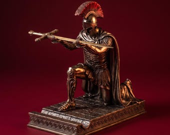 May include: A bronze-colored statue of a kneeling warrior wearing a plumed helmet and armor, holding a long sword horizontally with both hands. The figure is positioned on a rectangular base decorated with a repeating Greek key pattern.