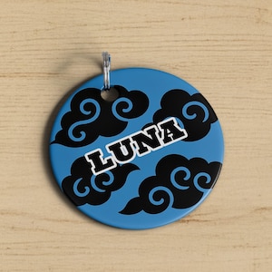 May include: A round, blue tag with the name "LUNA" in white, outlined in black. The tag features black cloud designs. A silver-colored metal loop is attached at the top for hanging.