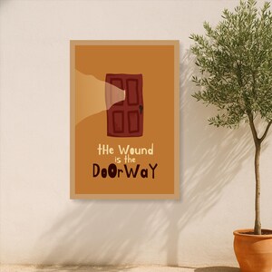 May include: A framed art print featuring a stylized illustration of a maroon door with a light shining through it. The text "The Wound is the DoorWay" is below the door. The artwork is set against a tan background.