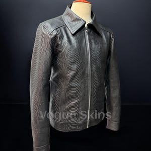 May include: A black, textured leather jacket with a full zipper closure and a collared neckline. The jacket is displayed on a mannequin with a red top. The jacket has a reptile skin pattern. The text "Vogue Skins" is visible on the jacket.