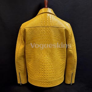 Może przedstawiać: Żółta skórzana kurtka o fakturze skóry krokodyla. Kurtka ma kołnierz, kieszenie zapinane na zamek i zapięcie na zamek. Tekst "Vogueskins" jest widoczny z tyłu kurtki.