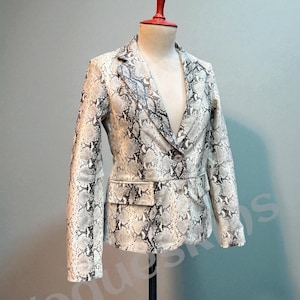 May include: A snakeskin-patterned blazer displayed on a mannequin. The jacket features a notched lapel, two front pockets, and a single-button closure. The print is a mix of gray, black, and cream tones. The mannequin has a red top.