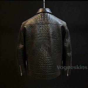Puede incluir: Chaqueta de cuero negro con textura de piel de cocodrilo, colgada de una percha. La chaqueta tiene un cuello clásico, bolsillos con cremallera y un patrón texturizado. La marca "Vogueskins" es visible en la parte inferior derecha.