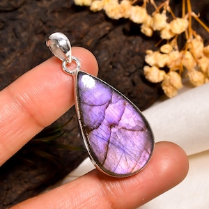 Könnte beinhalten: Ein tropfenförmiger Anhänger mit einem leuchtenden lila Edelstein. Der Stein hat ein einzigartiges, strukturiertes Aussehen mit Lavendel- und Violetttönen. Der Anhänger ist in einen silberfarbenen Metallrahmen gefasst und hat eine kleine Schlaufe.