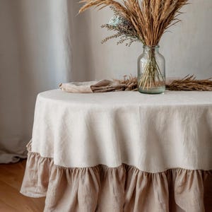 May include: A round, cream linen tablecloth with a tan ruffled edge. A clear glass vase displays dried wheat and grasses. A folded linen napkin sits on the table. The table is set for a rustic, natural aesthetic.