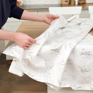 Peut inclure: Une nappe blanche ornée d'un motif floral délicat de brins gris et roses. Le tissu est déplié sur une table en bois, mettant en valeur le motif. La nappe est de couleur neutre et douce.