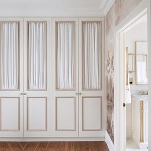 Pode incluir: Quatro portas de armário brancas com molduras bege e cortinas de tecido branco. As portas têm pequenos puxadores redondos prateados. O quarto tem chão de madeira e uma vista de uma casa de banho com acessórios dourados.