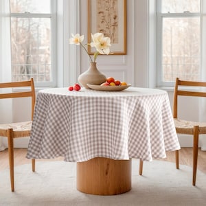 May include: A round table with a light brown wooden base is covered with a beige and white checked tablecloth. A vase with flowers and a bowl of fruit sit on the table. Two wooden chairs flank the table.