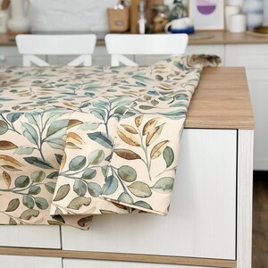 Peut inclure: Une nappe beige avec un motif botanique à l'aquarelle représentant des feuilles vertes, bleues et marron. Le tissu est posé sur un comptoir en bois, mettant en valeur le motif de feuilles et les variations de couleurs.