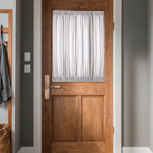 May include: A wooden door with a rectangular window covered by a white curtain. The door has a brass handle and is set in a white-trimmed frame. The door's wood grain is visible, and the surrounding walls are painted gray.
