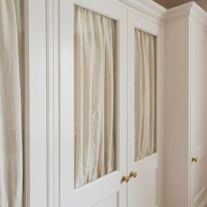 May include: White built-in cabinet with glass-paned doors. Cream-colored curtains hang behind the glass. The cabinet doors have brass knobs. The cabinet is part of a larger built-in unit.