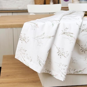 May include: White tablecloth with a delicate floral pattern featuring green and pink botanical elements. The fabric drapes over a wooden table, showcasing the design. The tablecloth is suitable for kitchen or dining room decor.