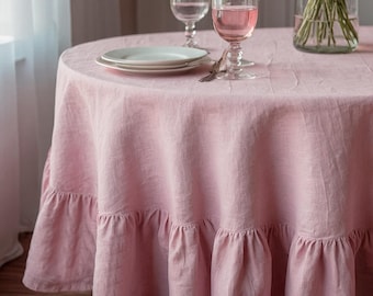 May include: A round, light pink linen tablecloth with a ruffled edge. Two white plates, two wine glasses, and a vase with greenery are on the table. The tablecloth is draped over a round table.
