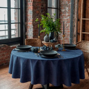 May include: A round dining table set for a meal, featuring a navy blue tablecloth. Black plates, silverware, and decorative gourds are arranged on the table. Tall black candles in gold holders and a vase with greenery complete the setting.