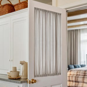 May include: A white interior door with a sheer, light gray curtain panel. The door has a brass doorknob and is slightly ajar, revealing a glimpse of a bedroom with a bed and curtains. Wicker baskets sit atop a white cabinet.