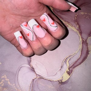 Könnte beinhalten: Eine Hand mit langen, quadratischen Kunstnägeln. Die Nägel sind mit einer hellrosa Basis, weißen Spitzen und einem silbernen Glitzerwellenmuster mit kleinen roten Herzen bemalt. Der Hintergrund ist rosa und lila marmoriert.