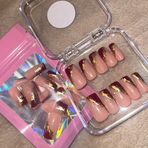 Könnte beinhalten: Ein Set künstlicher Nägel in einer transparenten Kunststoffbox und einem rosa Beutel. Die Nägel sind hellrosa mit einer dunkelroten Spitze und goldenen Folienakzenten. Die Box ist geöffnet und zeigt eine Reihe der Nägel.