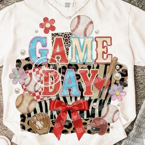 May include: White t-shirt with a baseball-themed design. The design includes the words "GAME DAY" in red and blue, baseballs, a baseball bat, and a red bow. The shirt has a crew neck and short sleeves.