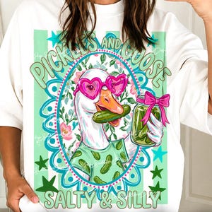 May include: White t-shirt with a whimsical graphic of a goose wearing heart-shaped sunglasses and holding a pickle and a jar of pickles. The text on the shirt reads "Pickles and Goose, Salty & Silly". The design includes green and pink accents.