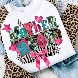 Könnte beinhalten: Weißes Sweatshirt mit dem Wort "MOTHERHOOD" in einem farbenfrohen Patchwork-Design. Die Buchstaben sind eine Mischung aus Pink, Türkis, Zebra- und Leopardenmuster. Eine rosa Schleife und Herzen akzentuieren das Design. Das Sweatshirt liegt auf blauen Jeans und Leopardenmuster-Stoff.