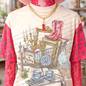 May include: Cream-colored t-shirt with a graphic of a shopping cart filled with vintage items, including a candelabra, picture frames, and blue and white china. The shirt has pink sleeves with cactus patterns and the text "I'd Rather Be Thrifting".