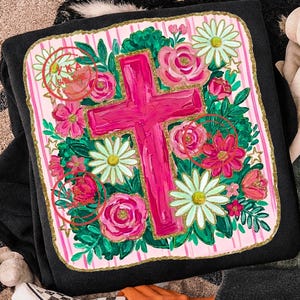 Peut inclure: Sweat-shirt noir orn&eacute; d'un motif floral vif avec une grande croix rose au centre. Le motif comprend des roses roses, des marguerites et du feuillage vert, le tout sur un fond &agrave; rayures roses et blanches avec une bordure dor&eacute;e.