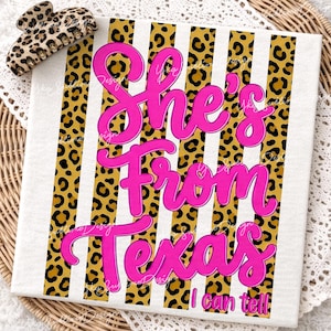 May include: A white canvas with the text "She's From Texas I can tell" in bright pink script. The background features alternating vertical stripes of leopard print and white. A leopard print hair clip rests on the top left.
