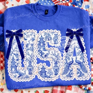 May include: A royal blue sweatshirt featuring the word "USA" in a white and blue chinoiserie pattern. The letters are edged with a scalloped trim and embellished with navy blue velvet bows. The shirt is decorated with the text "Yka Sublime Design".
