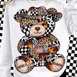 Pode incluir: Camisola branca com um design de urso de peluche em patchwork. O urso tem um chapéu de cowboy e um coração com a palavra "LOVE". As aplicações incluem estampado de leopardo, chamas e as palavras "HOWDY" e "VALENTINE".