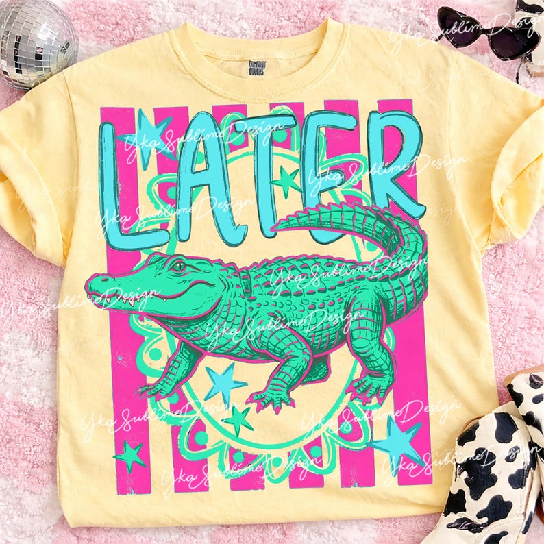 May include: Yellow t-shirt with a teal and pink graphic. The graphic features the word "LATER" in teal, above a green alligator. The alligator is surrounded by pink stripes and teal stars.
