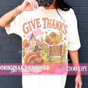 May include: An off-white t-shirt featuring a colorful graphic with the text GIVE THANKS and EST. 1621. The illustration includes a turkey wearing a pilgrim hat, a cornucopia filled with autumn produce, a large house, and several pumpkins.
